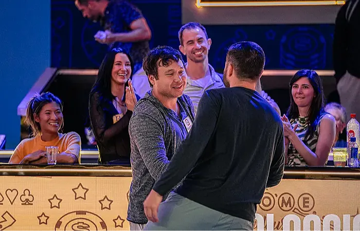 James Carroll e Jesse Yaginuma se cumprimentam e sorriem após uma partida de pôquer, enquanto um grupo de pessoas ao fundo aplaude e celebra. A cena acontece em um ambiente descontraído e iluminado, com uma bancada decorada com símbolos de pôquer como paus, ouros, copas e espadas. - rakebackpokerworld
